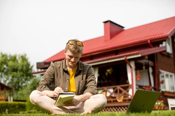 Joven descalzo sentado sobre hierba verde tomando notas en su cuaderno — Foto de Stock