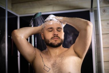 Genç, güzel adam banyo yapıyor, dövmeli yakışıklı adam saçlarını şampuanla yıkıyor.