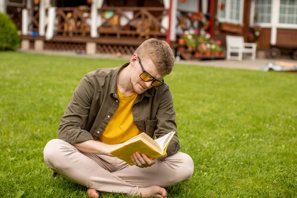 Sarı gözlüklü genç adam yeşil çimlerin üzerinde oturmuş kitap okuyor. — Stok fotoğraf
