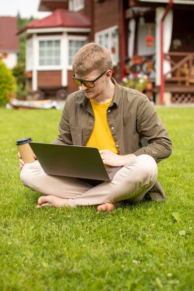 Joven hombre de negocios guapo trabaja en el ordenador portátil al aire libre, se sienta con café en la hierba — Foto de Stock