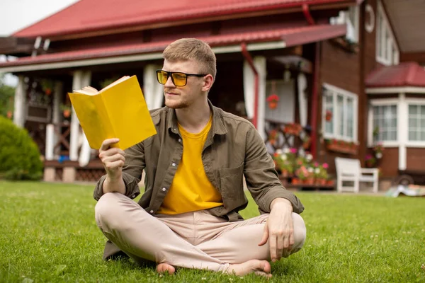 Genç ve zeki bir adam doğada dinleniyor kitap okuyor dışarıda çimlerin üzerinde oturuyor. — Stok fotoğraf