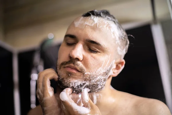 young bearded naked brunette man washes his beard with shampoo foam