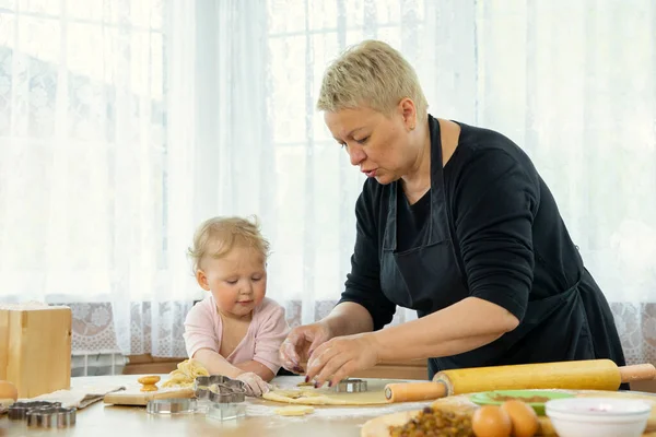 Grandmother in black apron teaches her cute little granddaughter make cookies