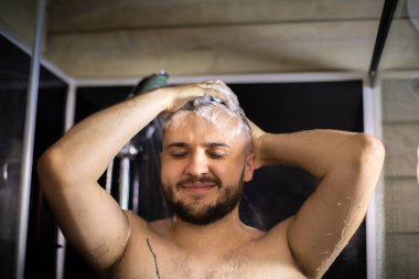 Genç, sakallı, çıplak esmer adam duşta başını şampuan köpüğüyle yıkıyor.