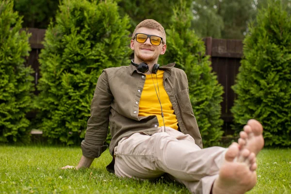 Descalzo joven se sienta en la hierba verde con auriculares en el cuello —  Fotos de Stock
