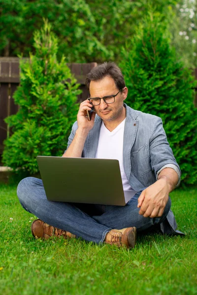 Hombre de negocios de mediana edad se sienta en la hierba trabaja en el ordenador portátil, habla en el teléfono móvil — Foto de Stock