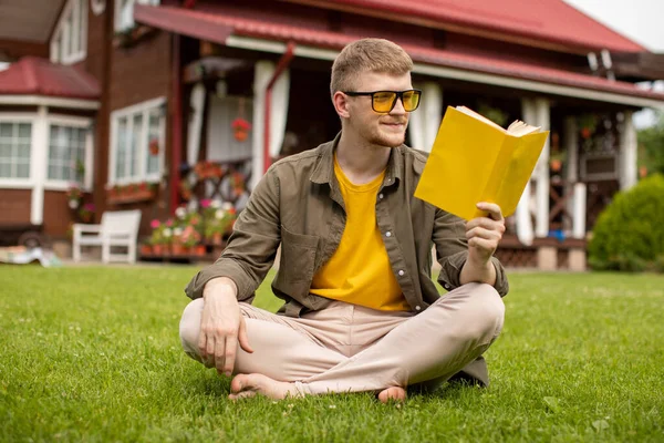 Sarı gözlüklü çıplak ayaklı genç adam yeşil çimlerin üzerinde oturmuş kitap okuyor. — Stok fotoğraf