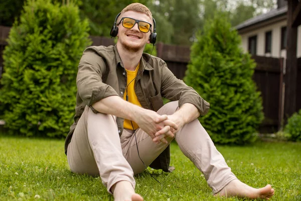 Descalzo joven se sienta sobre hierba verde escuchando música en los auriculares —  Fotos de Stock