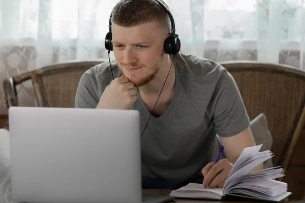 Joven en auriculares trabaja con el ordenador portátil haciendo notas a diario en la casa de madera — Foto de Stock