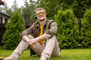 Kulaklıklı genç yakışıklı adam çimlerin üzerinde oturmuş eğitici podcast dinliyor.