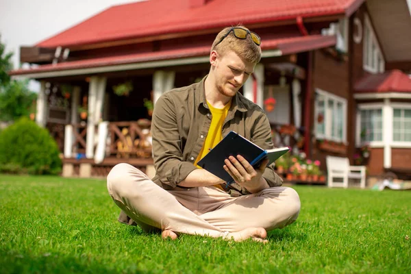 Yeşil çimlerin üzerinde oturan yalın ayak genç adam defterine notlar alıyor. — Stok fotoğraf