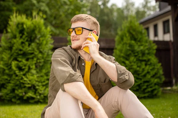 Joven hombre sonriente en gafas amarillas sentado en la hierba verde hablando en el teléfono inteligente — Foto de Stock