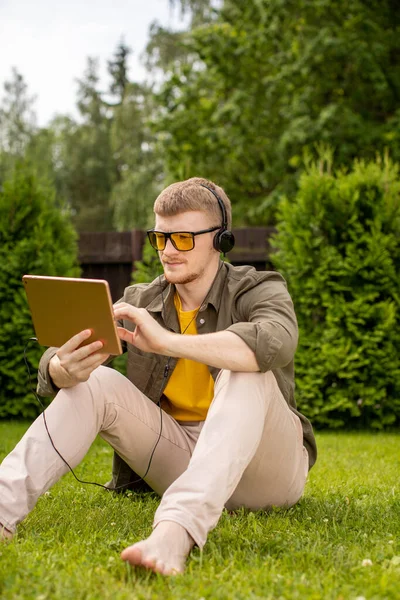 Descalzo joven sentado en la hierba verde con la tableta escuchando música — Foto de Stock