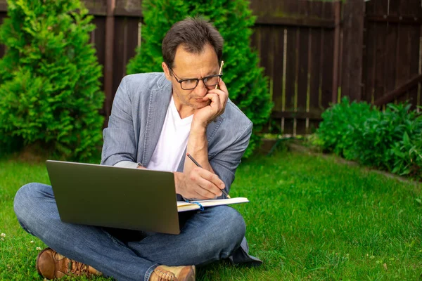 Brooding empresário raspado senta-se na grama com laptop fazendo anotações no notebook — Fotografia de Stock