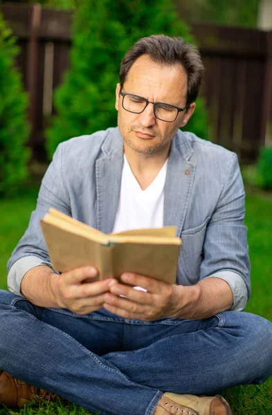 Sakin, traşsız, gözlüklü bir adam çimenlerde oturur ve yaz günü açık havada kitap okur. — Stok fotoğraf
