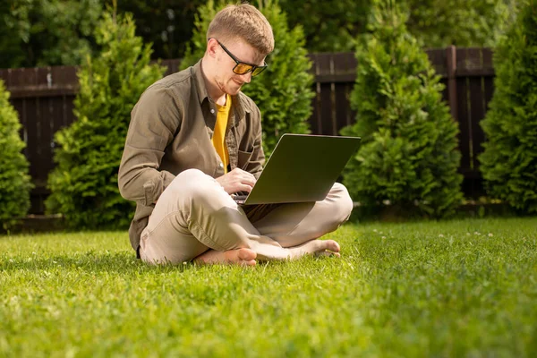 Joven hipster chico se sienta en la hierba en el parque de trabajo en el ordenador portátil, comprueba el correo electrónico — Foto de Stock