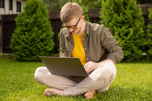 Joven hombre feliz descansando sentado en la hierba en el parque con el ordenador portátil en las manos — Foto de Stock