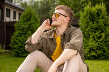 Sarı gözlüklü genç adam yeşil çimenlerde oturmuş akıllı telefonuyla konuşuyor.