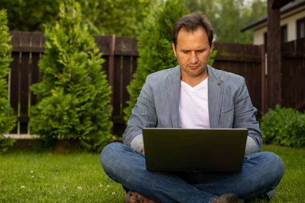 Distance work concept. 40s businessman sits crossed legs on grass with laptop