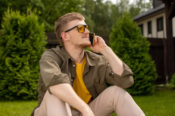 Lindo joven mira hacia otro lado hablando en el teléfono móvil mientras está sentado solo en la hierba — Foto de Stock