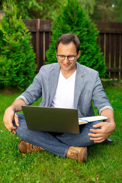 Alegre sonriente sin afeitar 40 empresario se sienta con las piernas cruzadas en la hierba con el ordenador portátil — Foto de Stock