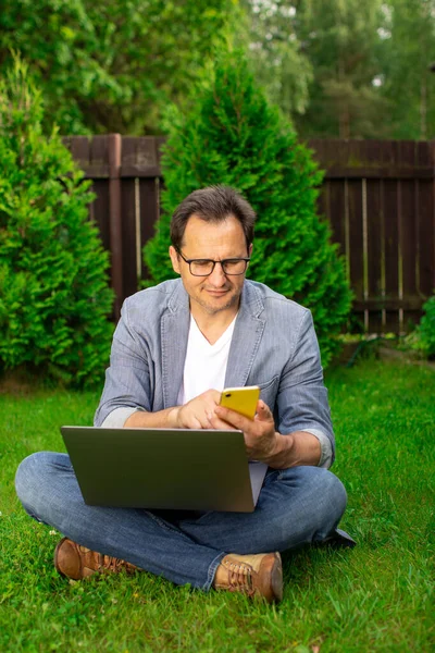 Tranquilo hombre de negocios de 40 años seguro se sienta en la hierba con el ordenador portátil escribiendo en el teléfono inteligente — Foto de Stock