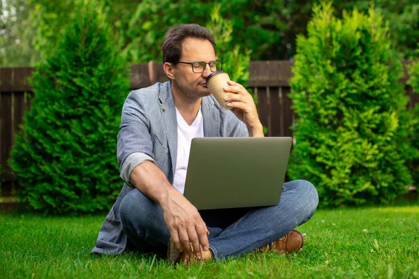 Calma positiva madura 40 hombre de negocios se sienta en la hierba con el ordenador portátil beber café — Foto de Stock