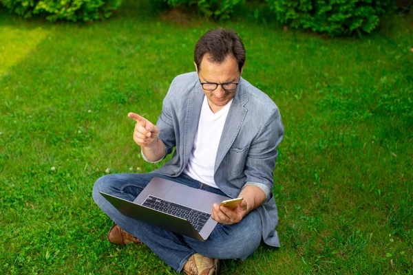 Alegre centrado hombre de negocios sin afeitar se sienta en la hierba con el ordenador portátil señalando el dedo — Foto de Stock