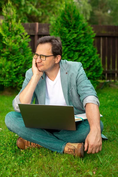 Adulto hombre pensativo se sienta al aire libre con el ordenador portátil y portátil mira hacia otro lado, freelancer — Foto de Stock