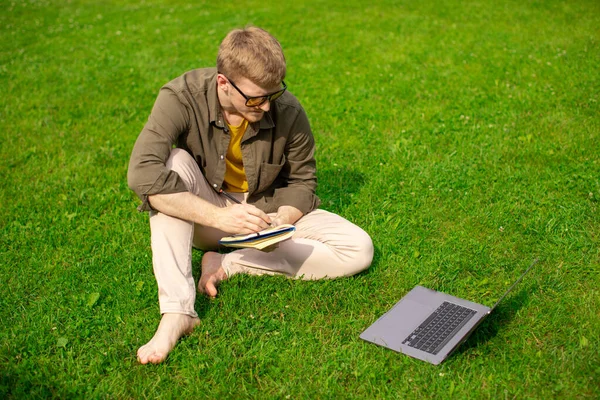 Joven millenial chico se sienta en la hierba viendo webinar distancia en línea, toma notas — Foto de Stock