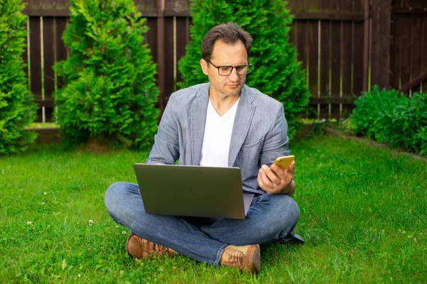 Tranquilo hombre de negocios de 40 años seguro se sienta en la hierba con el ordenador portátil mirando el teléfono inteligente — Foto de Stock