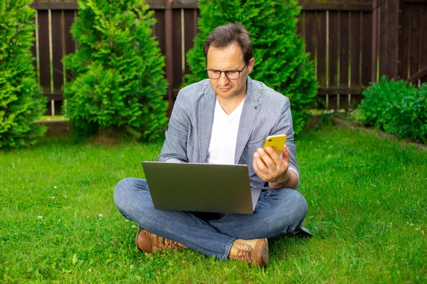 Hombre adulto sentado en el césped al aire libre trabajando en el ordenador portátil, escribiendo en el teléfono móvil — Foto de Stock