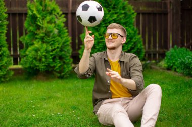 Mutlu gülümseyen beyaz bir adam yeşil zeminde futbol topunu parmağında döndürür.