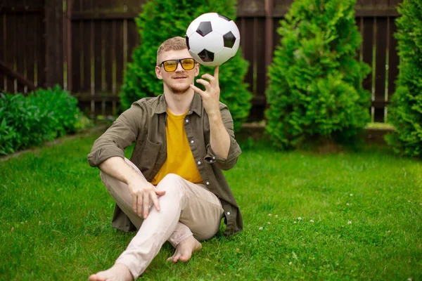 young man in everyday clothes spins soccer ball on finger on green lawn