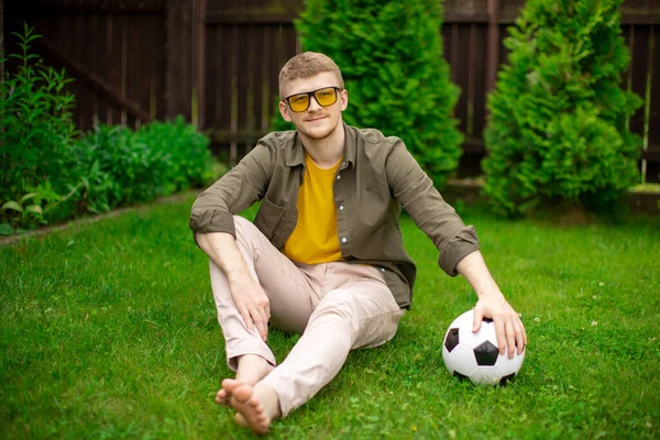 Joven hipster chico se sienta en la hierba con pelota de fútbol, ventilador de fútbol tienen tiempo libre —  Fotos de Stock