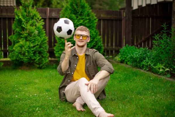 Hombre guapo se sienta en el césped haciendo girar la pelota de fútbol en el dedo índice, apuestas deportivas —  Fotos de Stock