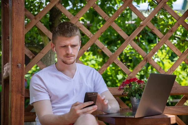 Hombre joven que trabaja en el ordenador portátil escribiendo en el teléfono móvil en la mañana de verano — Foto de Stock