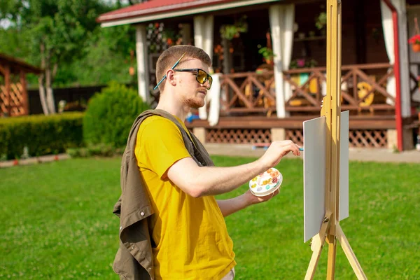 Joven hombre atractivo en gafas de sol de moda pintura de imagen en el caballete exterior — Foto de Stock