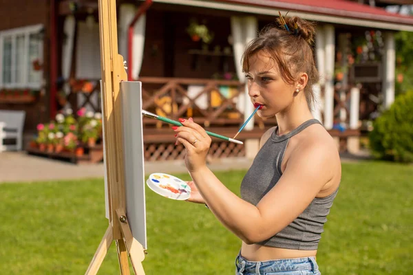 Young beautiful female artist paints on easel outside holds paint brush in mouth