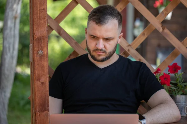 Ung man bär svart t-shirt tittar på laptop på trä pergola rutnät bakgrund — Stockfoto