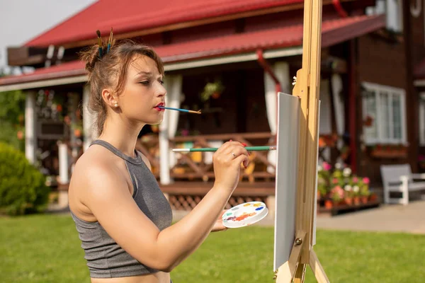 Όμορφο κορίτσι ζωγραφίζει easel ζωγραφική κρατώντας βούρτσα στα δόντια έξω από το σπίτι χώρα — Φωτογραφία Αρχείου