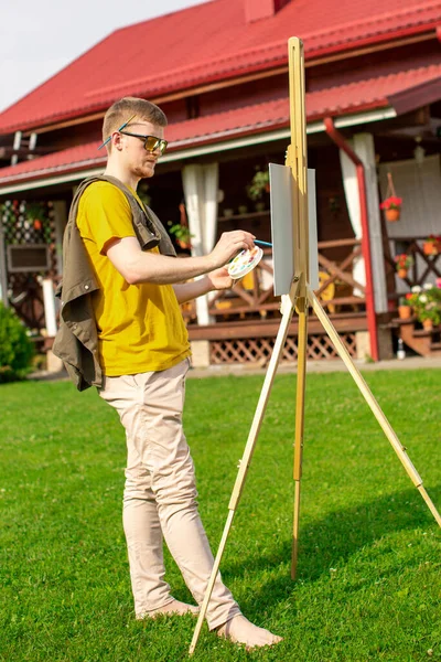 Young attractive male artist painting on easel outside on loan near countryhouse — Stock Photo, Image