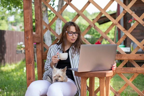 young woman works with laptop drinking coffee on backdrop of wooden pergola grid