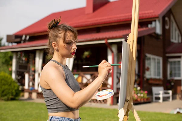 Joven hermosa artista femenina pinta en caballete fuera en préstamo cerca de casa de campo — Foto de Stock