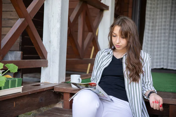 Chica morena elegante de moda leyendo revista en la terraza de la casa de madera — Foto de Stock