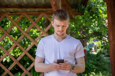 Düşünceli sakallı yakışıklı genç adam açık havada akıllı telefona bakıyor.