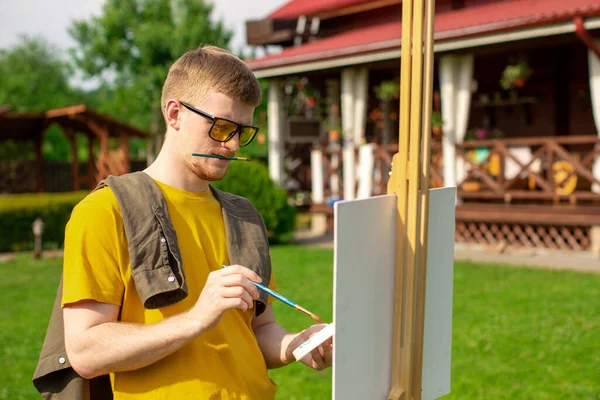 Νέος δημιουργικός καλλιτέχνης σε μοντέρνα κίτρινα γυαλιά ηλίου ζωγραφίζει σε easel έξω — Φωτογραφία Αρχείου