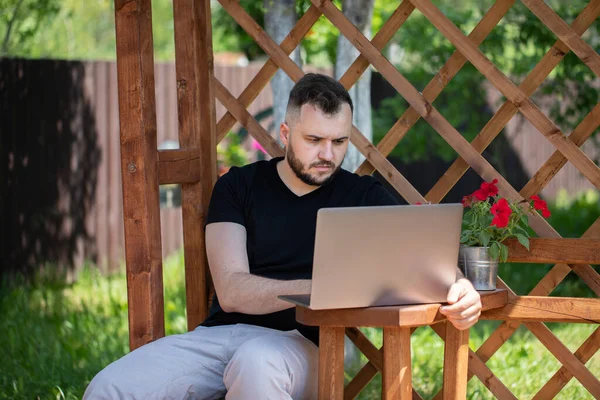 Guapo joven trabaja con el ordenador portátil al aire libre en pérgola de madera en verano — Foto de Stock