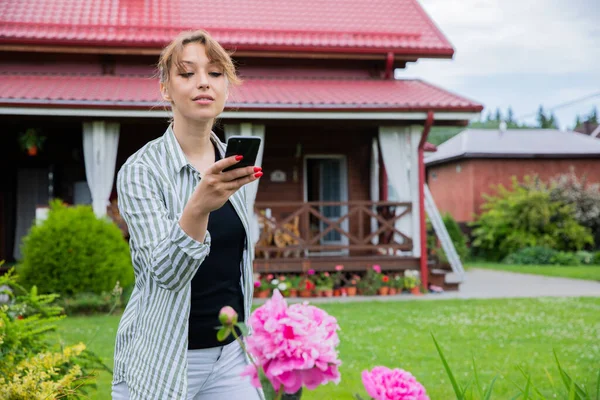 joyful young brunette woman takes pictures of pink peony flowers in her garden