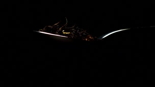 Schwarzer Blatttee mit getrockneten Blütenblättern, die aus einem Teelöffel auf Schwarz fallen — Stockvideo